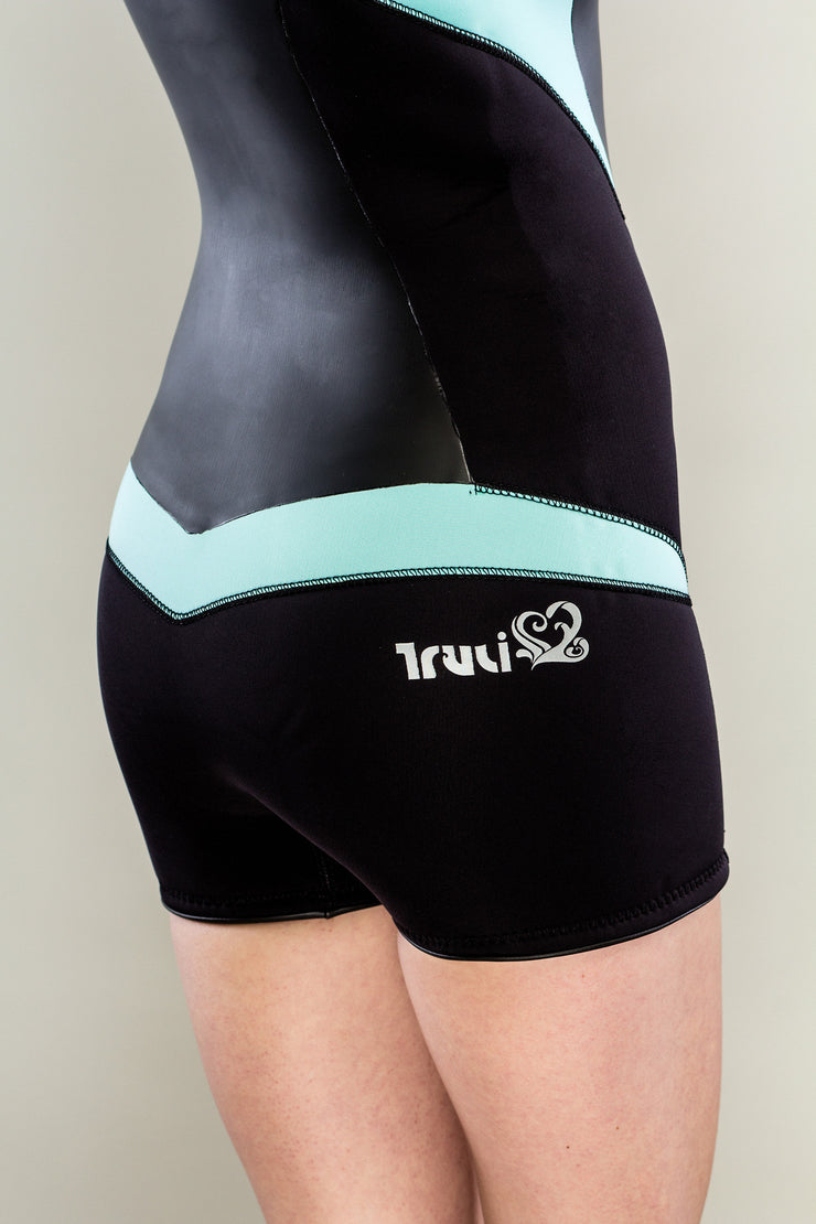 A slender woman is standing in a sleeveless shorty Truli-Mi wetsuit by Truli Wetsuits for women. The trim is light blue and the rest of the wetsuit is black. The view is from her backside up to her chest only.